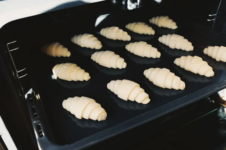 Close-up of raw croissants baking on a tray inside an oven, showcasing homemade pastry preparation.